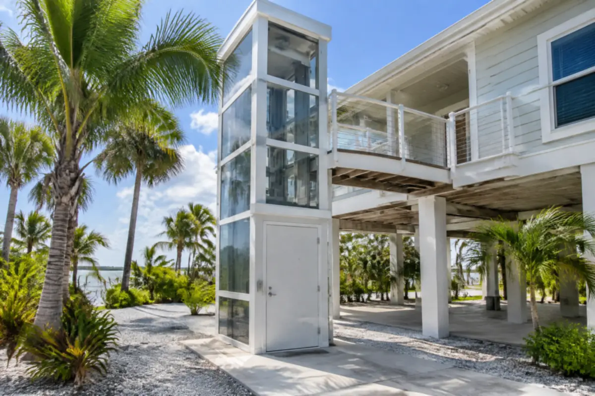 Glass and aluminium outdoor elevator tower beside a raised stilt home, with a solid door at ground level